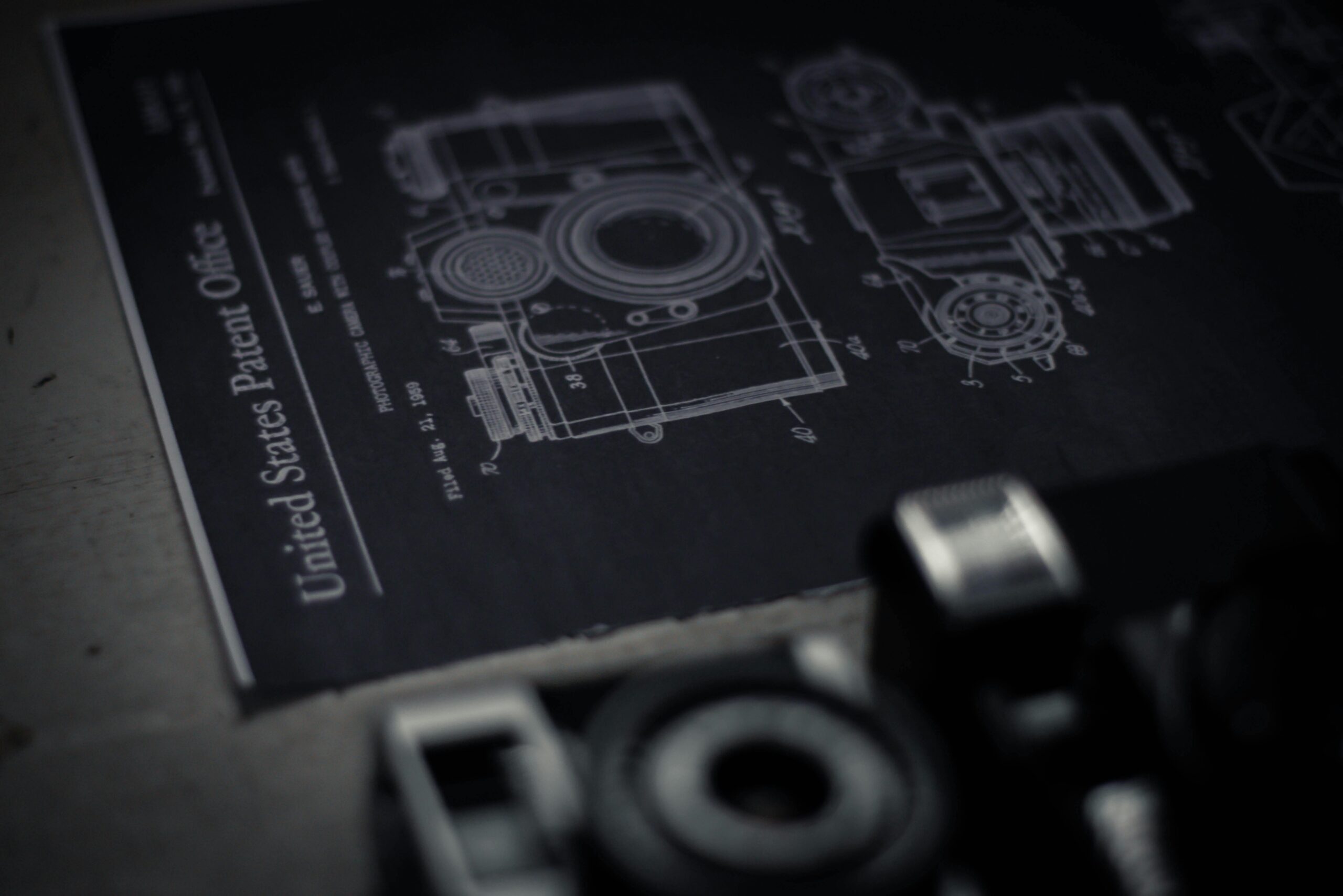 A dark, close-up image of a blueprint with out-of-focus screws beside it, symbolizing invention and patent complexity