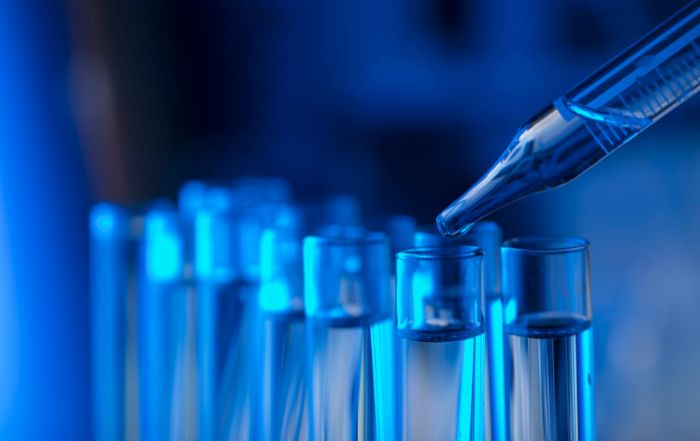 Dark blue-lit science test tube with substance dropping, illustrating PFAS chemical testing and regulation uncertainty
